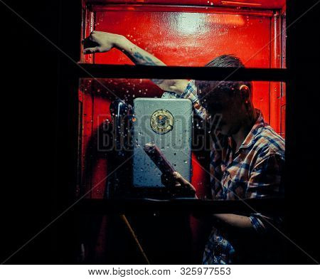 A Man Of Criminal Appearance In A Payphone Booth At Night. Talking On The Phone With A Revolver And