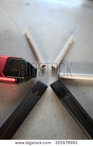 3 Vape Electronic Cigarette Devices And 3 Cigarettes In A Star Pattern On A White Textured Backgroun