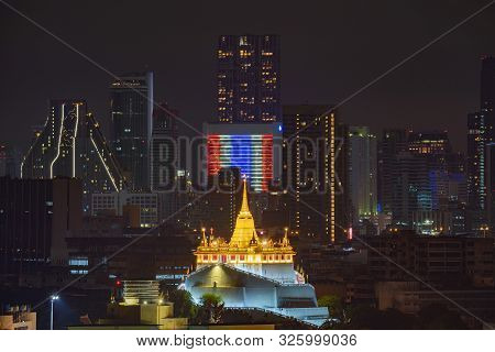 Golden Mount Or Wat Saket, Phukhao Thong, And Skyscraper Buildings With Thailand Flag At Night In Ba
