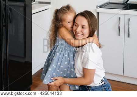 Child Whispering Something On Mothers Ear At Home On The Kitchen. Kid Telling A Secret. Toddler Girl