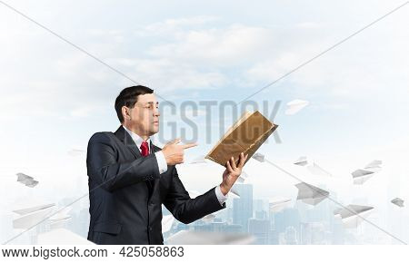 Senior Businessman Finger Pointing Into Open Book. Man In Business Suit And Tie Standing On Cityscap