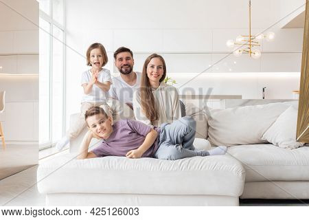 Family Portrait Of Young Parents With Their Two Children At Home, White Modern Desgin Of Living Room