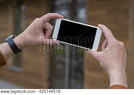 Man Holds A Mock-up Of A Smartphone With A Black Screen In His Hands On A Wooden Background.