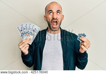 Young hispanic man playing poker holding dollars banknotes and chips angry and mad screaming frustrated and furious, shouting with anger looking up. 