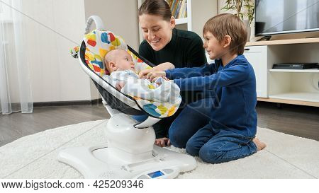 Older Brother And Smiling Mother Playing With Little Baby Boy Rocking In Electric Rocking Seat. Chil