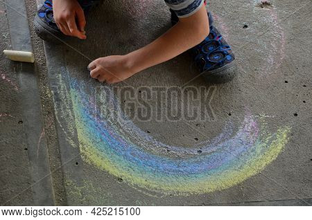Little Boy Draws A Rainbow On The Ground With Chalk. Holding A Pastel In His Hand And Drawing A Arc 