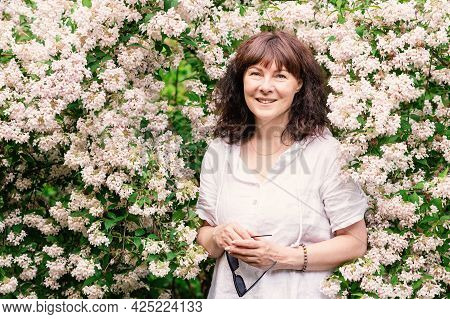 Portrait Nice Woman Of Forty Five Years Old With Brown Hair. Stands Near Large Flowering Bush, Smili