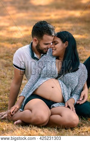 Young Couple Sitting On The Grass. The Young Woman Is Pregnant