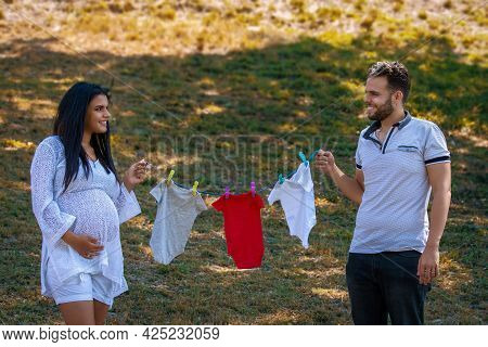 Young Couple Holding A Twine With Hanging Baby Clothes. The Young Woman Is Pregnant