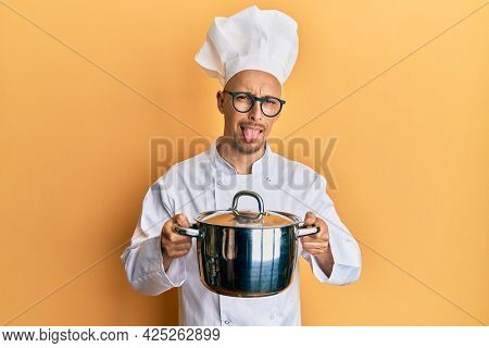 Bald man with beard wearing professional cook holding cooking pot sticking tongue out happy with funny expression. 