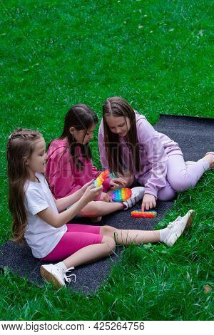 Three Teenage Girls In Colorful Clothes Play With Trendy Sensory Toy Pop It Outdors. Rainbow Soft Si
