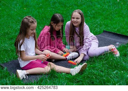 Three Teenage Girls In Colorful Clothes Play With Trendy Sensory Toy Pop It Outdors. Rainbow Soft Si