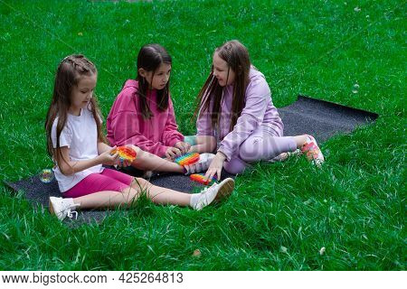 Three Teenage Girls In Colorful Clothes Play With Trendy Sensory Toy Pop It Outdors. Rainbow Soft Si