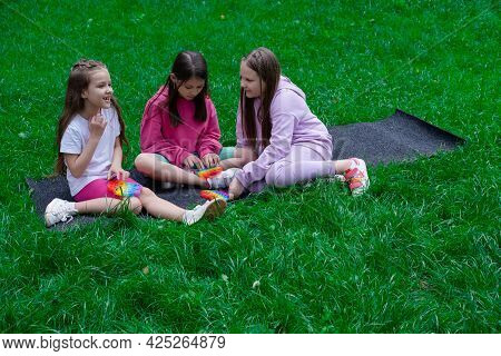 Three Teenage Girls In Colorful Clothes Play With Trendy Sensory Toy Pop It Outdors. Rainbow Soft Si