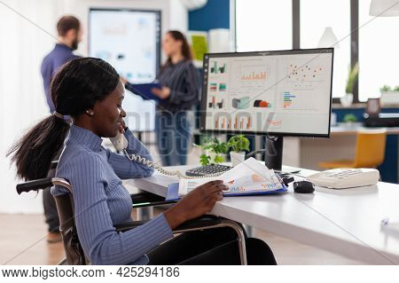 Busy Paralysed Handicapped Black Employee Answering Phone Talking With Colleague Sitting Immobilized