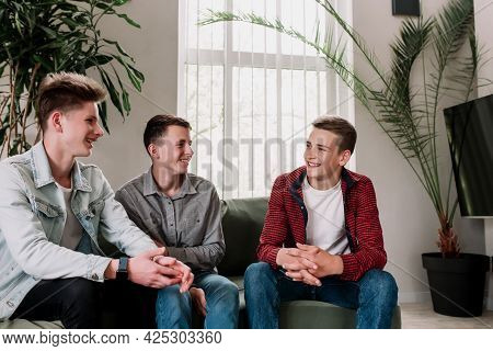 Three Male Friends Are Talking At The Green Sofa In Living Room. Guys Having A Talk, Sharing And Dis