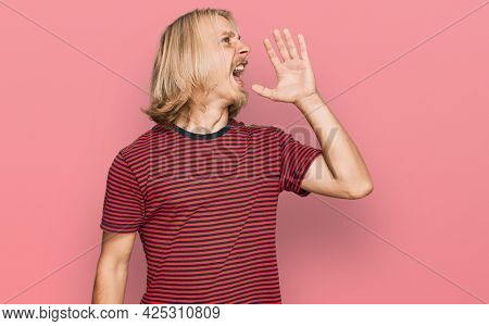 Caucasian man with blond long hair wearing casual striped t shirt shouting and screaming loud to side with hand on mouth. communication concept. 