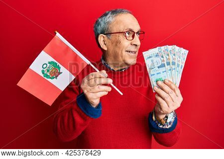 Handsome senior man with grey hair holding peru flag and peruvian sol banknotes smiling looking to the side and staring away thinking. 