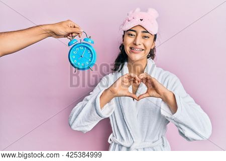Hispanic teenager girl with dental braces wearing sleep mask and robe with alarm clock close to her smiling in love doing heart symbol shape with hands. romantic concept. 