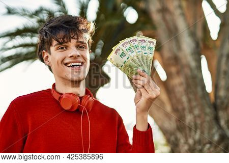 Young hispanic man using headphones holding argentinian pesos at the city