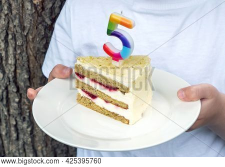 A Boy In A White T-shirt Holds A Piece Of Cake With A Candle On A Plate Against The Background Of Th