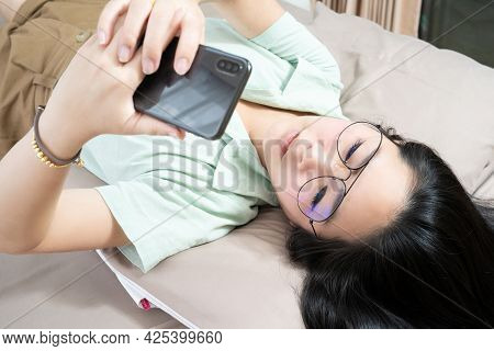 Beautiful Glasses Asian Woman Lie On Her Back, Chatting, Typing, And Texting, Mobile In Her Bedroom 