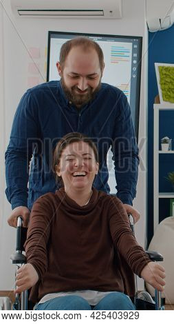 Cheerful Young Man Pushing Wheelchair With His Disabled Paralysed  Coworker While Smiling In 