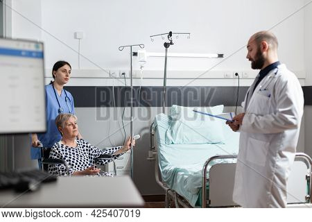 Senior Woman Getting Medicine Through Intravenous Line Bag Sitting On Wheechair Pushed By Nurse In H