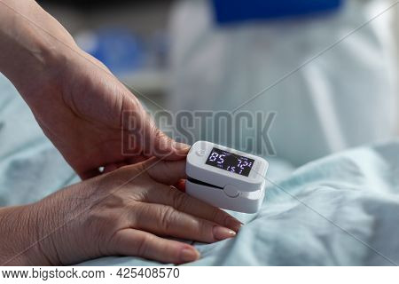 Close Up Of Oxymeter Attached On Sick Senior Patient Laying In Hospital Bed, Monitoring Hemoglobin. 