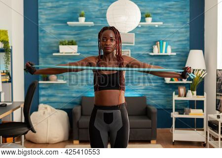 Black Strong Fit Woman Standing In Living Room Working Out Shoulders Using Resistance Band Dressed I