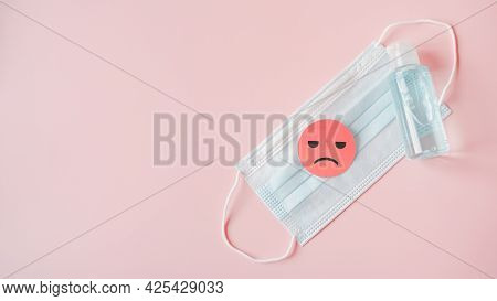 Unhappy Face On Pink Paper Cut On Medical Face Mask And Bottle Of Blue Alcohol Gel On Sweet Pink Bac