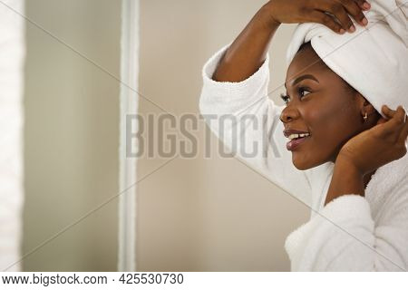 Smiling african american woman in bathroom wearing bathrobe, looking in mirror putting towel on head. health, beauty and wellbeing, spending quality time at home.