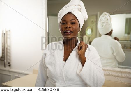 Portrait of happy african american woman in bathroom wearing bathrobe, with towel on head. health, beauty and wellbeing, spending quality time at home.