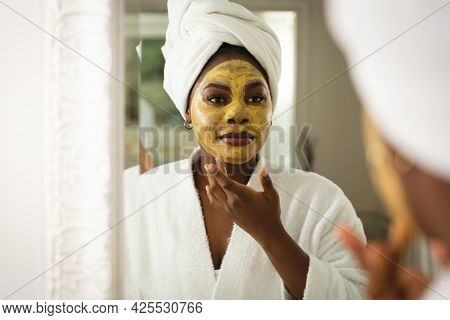 Happy african american woman in bathroom with towel on head, looking in mirror applying face mask. health, beauty and wellbeing, spending quality time at home.