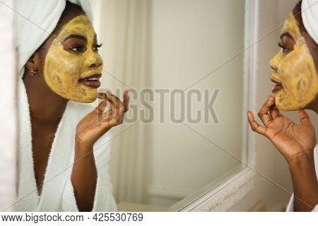 Happy african american woman in bathroom with towel on head, looking in mirror applying face mask. health, beauty and wellbeing, spending quality time at home.