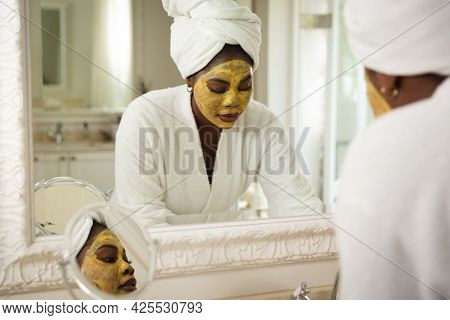 African american woman in bathroom with towel on head, wearing beauty treatment face mask. health, beauty and wellbeing, spending quality time at home.