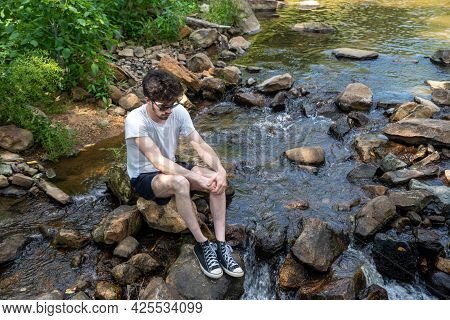 Caucasian Man Sits Alone In A Natural Setting Sitting On A Stepping Stone In The Middle Of A Stream.