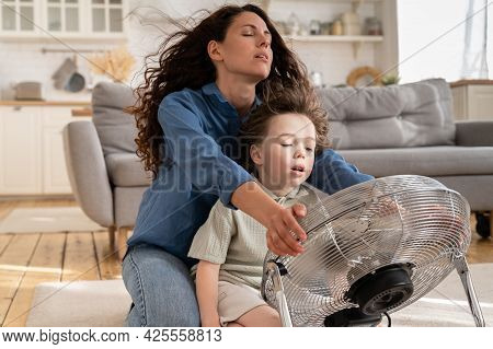 Happy Mom And Small Kid Refreshing Sit Near Big Indoor Ventilator Blowing Cooling Fresh Air At Home 