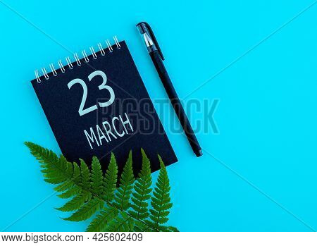 March 23th. Day 23 Of Month, Calendar Date. Black Notepad Sheet, Pen, Fern Twig, On A Blue Backgroun
