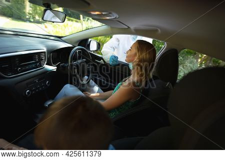 Medical worker wearing ppe suit taking swab tests of caucasian mother with son sitting in car. medical precautions, lifestyle during covid 19 pandemic.