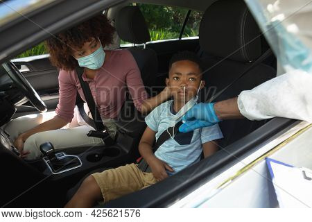 Medical worker wearing ppe suit taking swab tests of african american mother with son sitting in car. medical precautions, lifestyle during covid 19 pandemic.
