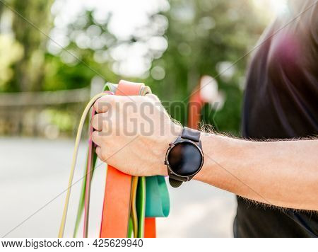 Strong man guy holding in his hand wearing fitness tracker set of colorful elastic rubber bands at the stadium outdoors. Athlete male person during workout with additional sport equipmant