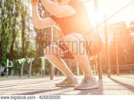 Strong sportive man guy doing squats with elastic rubber band at the stadium outdoors with sunlight. Adult male person during workout for legs with additional sport equipmant