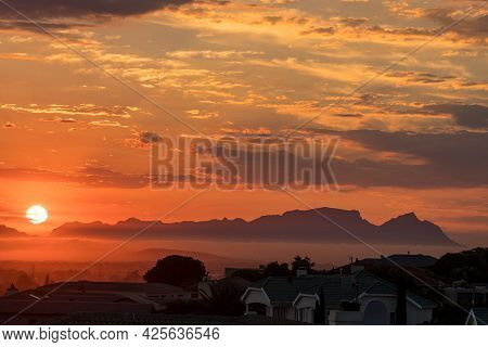 The Sun Is Setting Behind Fish Hoek Accross False Bay. The Back Of Table Mountain And Devils Peak Ar