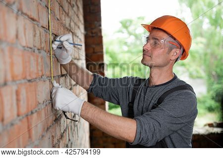 Construction Worker Male Foreman In Protective Helmet And Goggles Measures The Brick Wall Ruller. Re