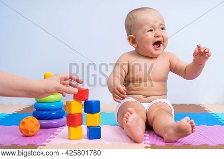 Crying Child In The Playroom. Mom Helps A Child Of 8-10 Months To Assemble A Construction Set From P