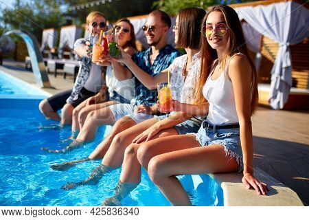 Group Of Friends Having Fun At Poolside Party Clinking Glasses With Fresh Cocktails Sitting By Swimm