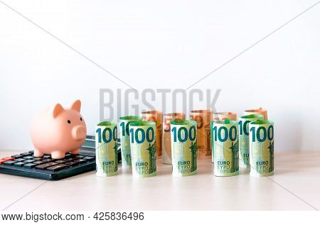 A Piggy Bank On A Calculator And Paper Money In Denominations Of 100 And 50 Euros, Folded Into A Rol
