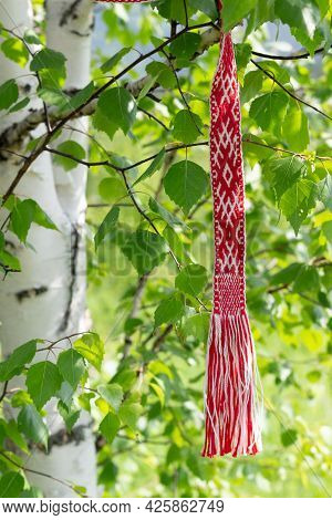 Red And White Ribbon With Ornament Of Traditional Slavic Signs Hanging On A Young Birch. Celebrating