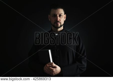 Priest In Cassock With Bible On Dark Background